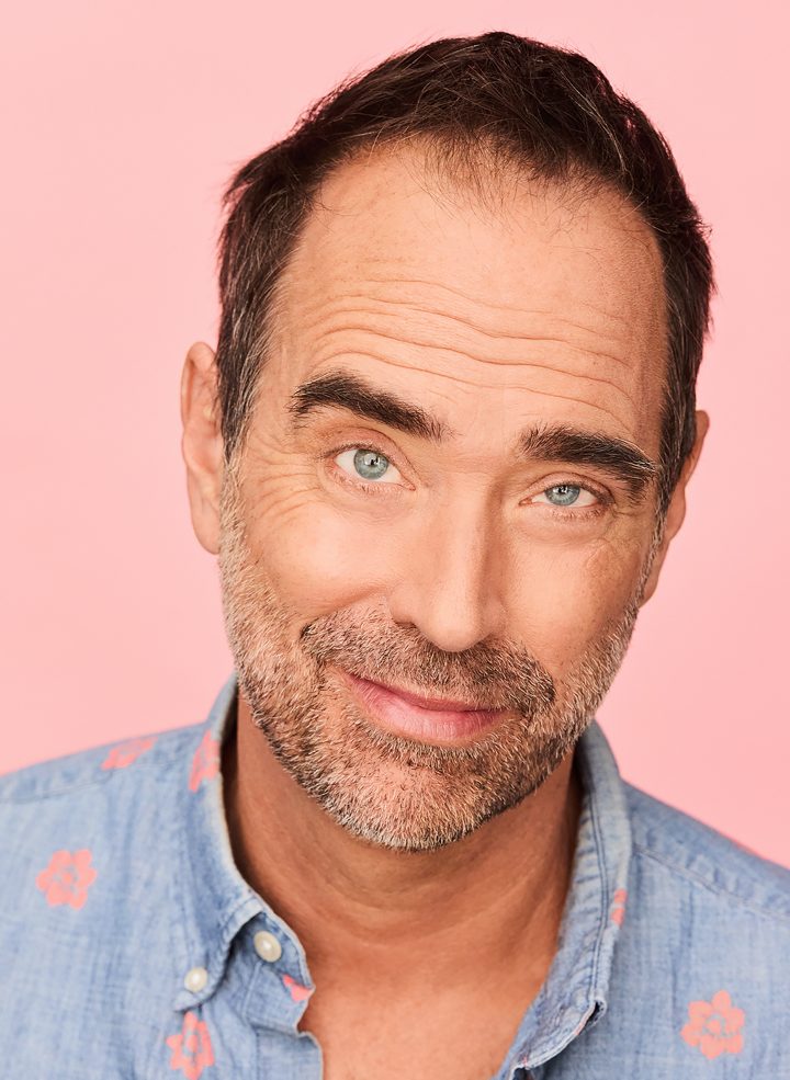 Headshot of Mike Romo: smiling with his head tilted, wearing a blue floral-print shirt against a pink background.